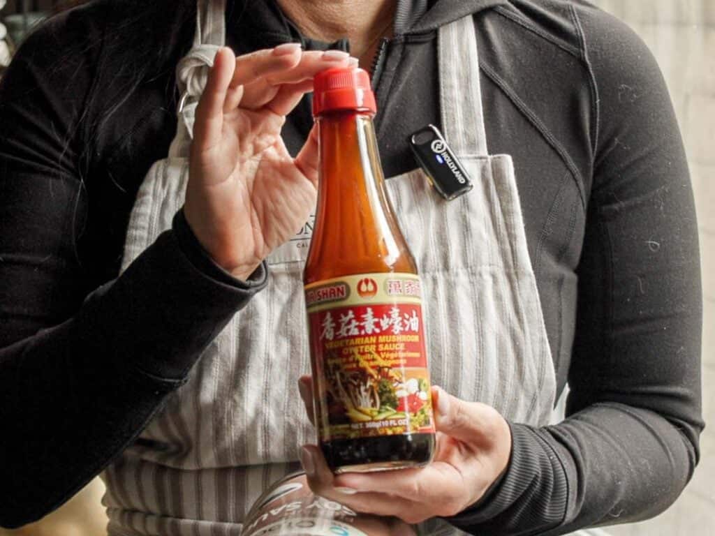 A person wearing a black shirt and striped apron holds a bottle labeled Vegetarian Mushroom Stir-Fry Sauce with red accents. The person’s face is not visible.